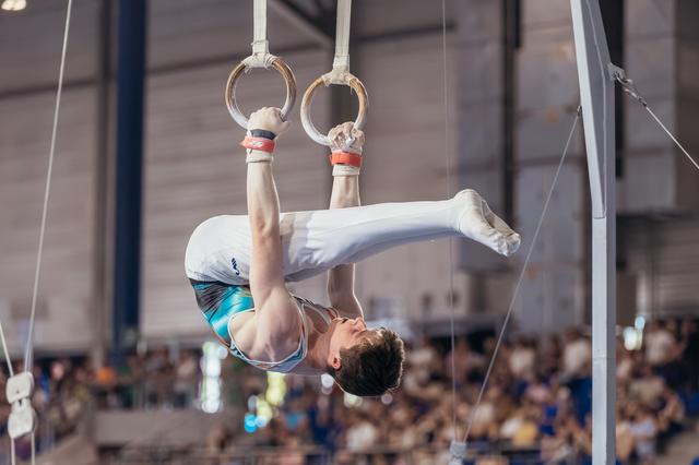 Young male gymnast hangs inverted on still rings, legs raised, showing intense concentration during a gymnastics meet.