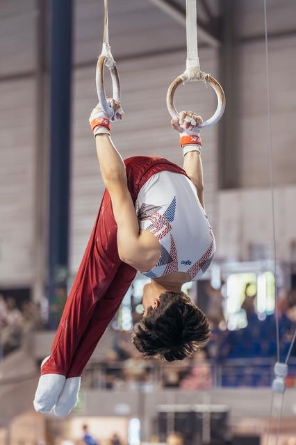 Young male gymnast performs an inverted hold on still rings, body curved with intense concentration in a competition hall.