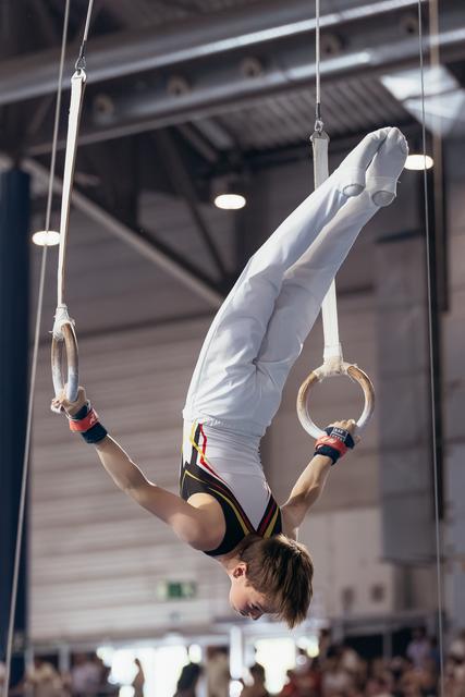 Young male gymnast performs an inverted pike hold on still rings, arms extended, body rigid with intense concentration.