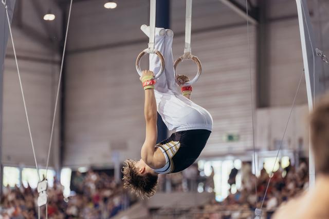 Young male gymnast performs an inverted hold on still rings, showing intense focus and strength in a packed arena.