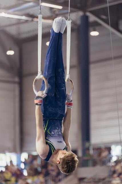 Young male gymnast performs an inverted hang on still rings, legs extended upward, showing intense concentration in a competition hall.