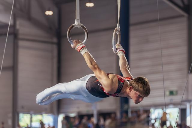 Young male gymnast holds an iron cross on still rings, body horizontal, intense concentration, indoor sports hall.