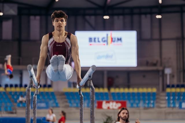 Young male gymnast holds an L-sit on parallel bars with intense focus, competing at a Belgium gymnastics event.