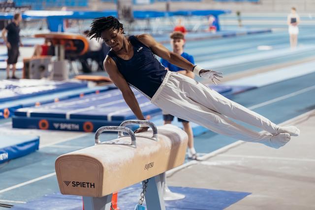 Young male gymnast performs a pommel horse routine, body fully extended in the air, intense focus on his face.