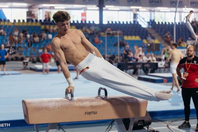 Young male gymnast performs on a Fujitsu pommel horse, body horizontal, expression focused during a competition.