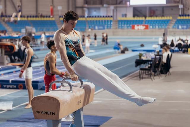 Young male gymnast performs on pommel horse, legs extended, concentrated expression in indoor arena.