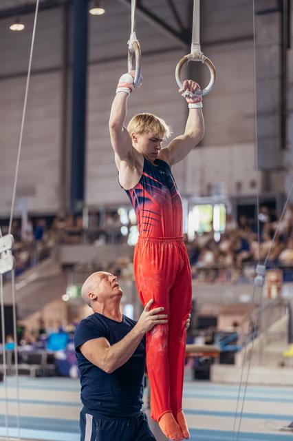 Young blond gymnast grips still rings with intense focus while coach spots his hips in a gymnasium.