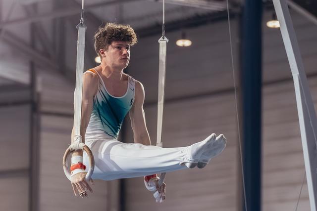 Young male gymnast performs an L-sit on still rings, his focused gaze and taut muscles showing intense concentration.