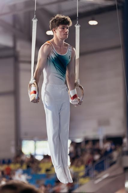 Young male gymnast hangs on still rings with intense focus, wearing teal leotard and white pants in a packed arena.