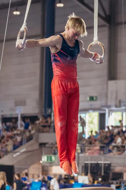 Blond male gymnast suspended on still rings, focused expression, wearing red and navy leotard in packed indoor arena.