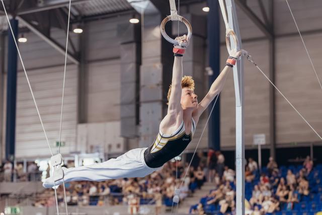 Young male gymnast performs a powerful horizontal hold on rings, body perfectly parallel, focused expression, in a packed arena.