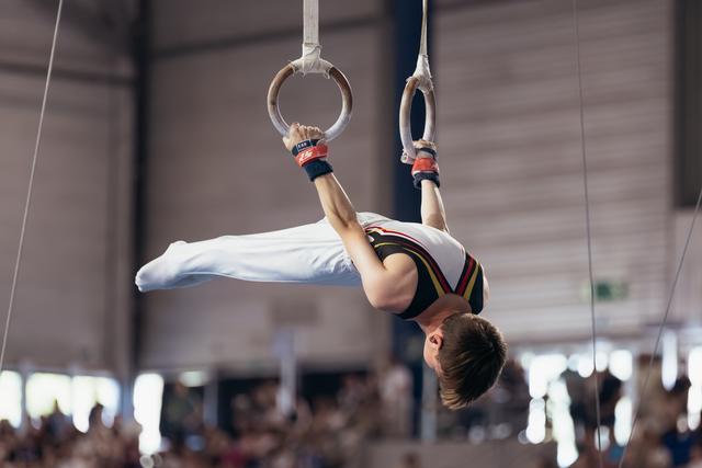 Male gymnast performs an inverted iron cross on still rings, body horizontal, intense focus during competition.