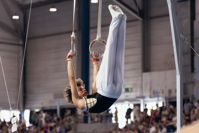 Young male gymnast performs an inverted pike hold on still rings, showing intense focus and strength before a crowd.