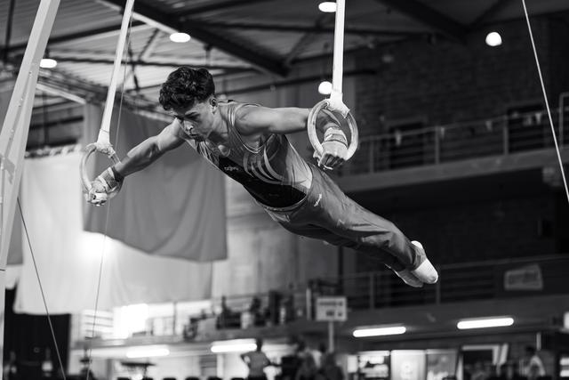 Young male gymnast holds an iron cross on still rings, body horizontal, expression intense with focused determination in indoor arena.