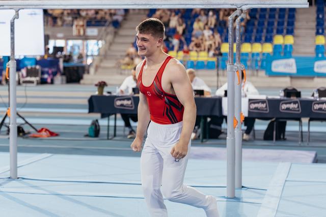 Young male gymnast in red leotard walks away from parallel bars, smiling proudly after his routine in a gymnastics arena.