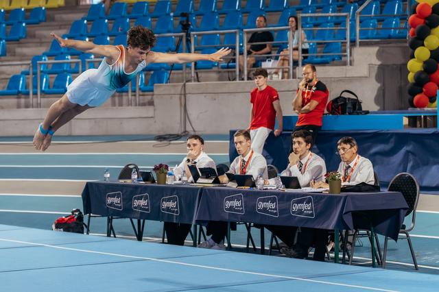 Male gymnast soars mid-air in a swan dive above judges at a Gymfed competition, arms outstretched with focus.