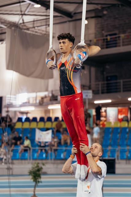 Young male gymnast holds still rings with intense focus while coach supports his legs in a competition gymnasium.