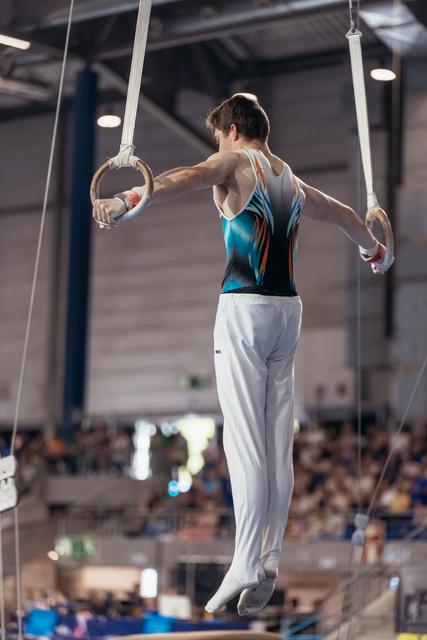 Male gymnast performs on still rings, suspended mid-air in a teal leotard, showing controlled strength before a crowd.