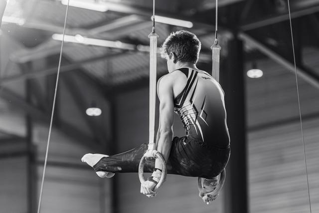 Male gymnast performs on still rings in a tuck position, showing focused strength and controlled technique in an indoor gymnasium.