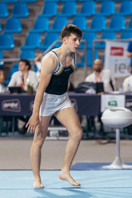 Young male gymnast in focused crouch position on floor exercise mat, wearing black Quatro leotard, in a competitive arena.