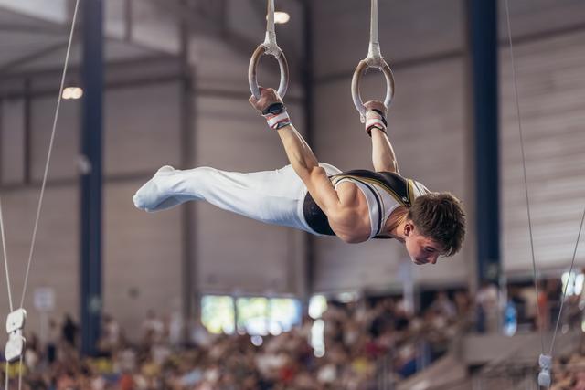 Young male gymnast performs an iron cross on still rings, body horizontal, intense focus, in a packed sports hall.