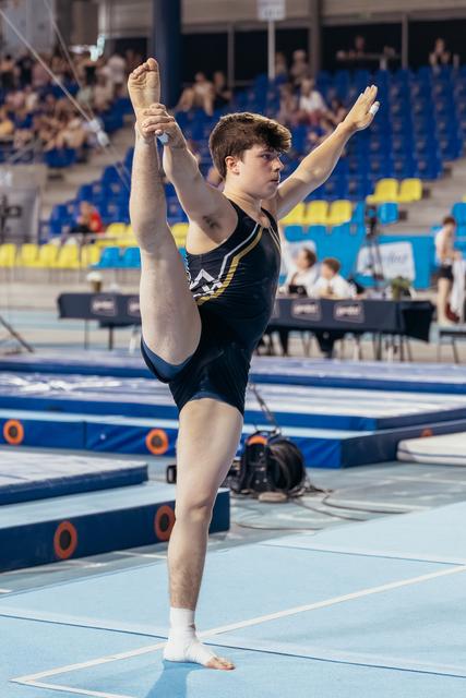 Young male gymnast performs a standing leg hold on the floor, focused expression, in a blue gymnastics arena.