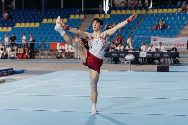 Young male gymnast performs a scale on floor, leg raised high, arms outstretched, focused expression in a blue-seated arena.