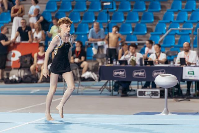 Young gymnast with red hair lands barefoot on floor mat, focused expression, judges table visible in background.