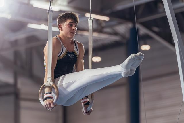 Young male gymnast performs on still rings with intense focus, legs raised, wearing white pants and dark vest in a gymnasium.