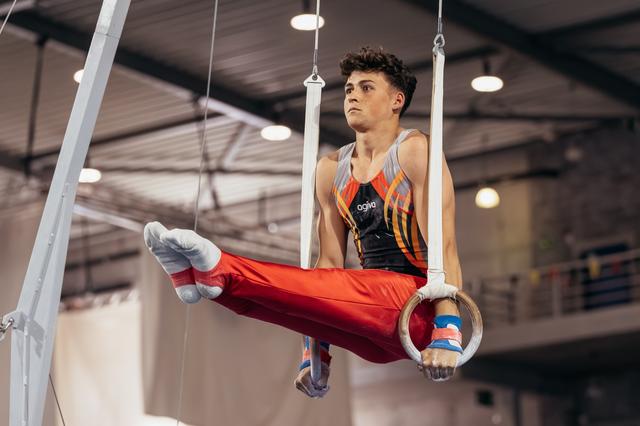 Young male gymnast holds an L-sit on still rings, wearing red pants and agiva kit, focused expression in indoor gym.