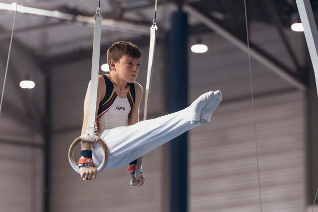 Focused young male gymnast holds still rings with legs raised, wearing agivo kit in a competition gymnasium.