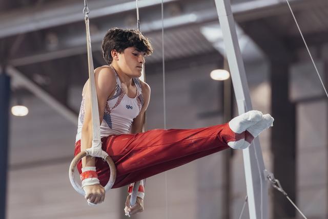 Young male gymnast holds a focused pike position on still rings, wearing red pants and white vest in a competition hall.
