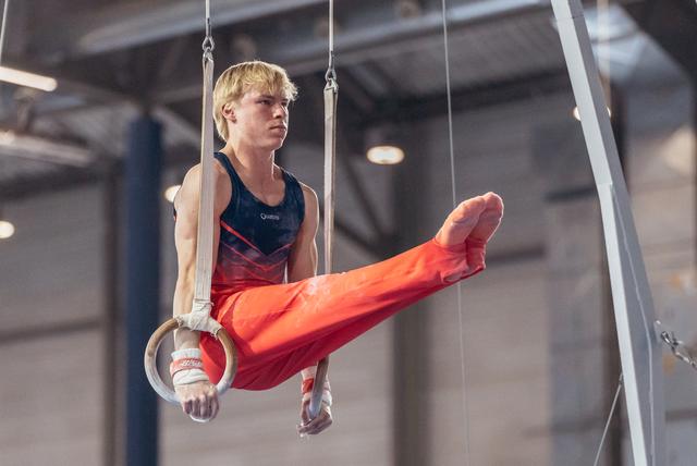 Focused young male gymnast swings on still rings, leg raised, wearing red and navy leo in a gymnasium.