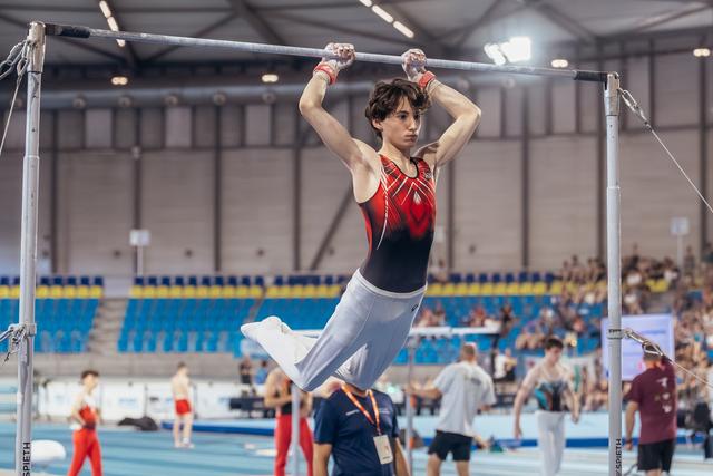 Young male gymnast hangs from the high bar with focused intensity, wearing red and black leo, in a large sports hall.
