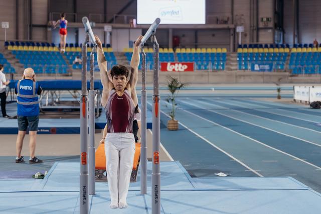 Young male gymnast hangs from parallel bars with focused expression, white pants and maroon leotard, indoor arena.