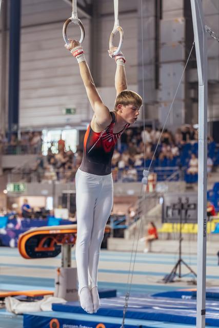 Young male gymnast hangs from still rings with focused expression, arms fully extended, in a busy sports hall.