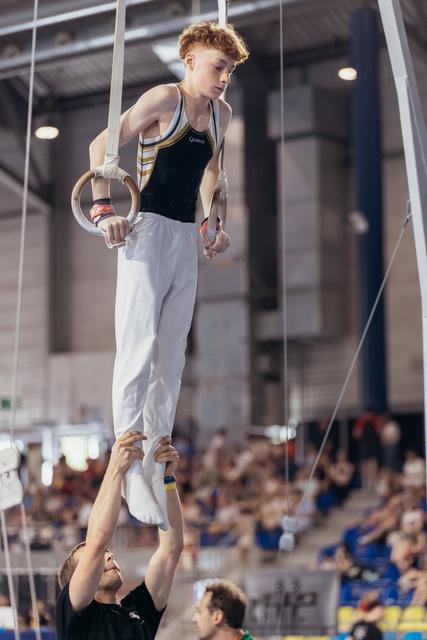 Young male gymnast grips still rings with focused concentration while coach steadies his legs in packed arena.