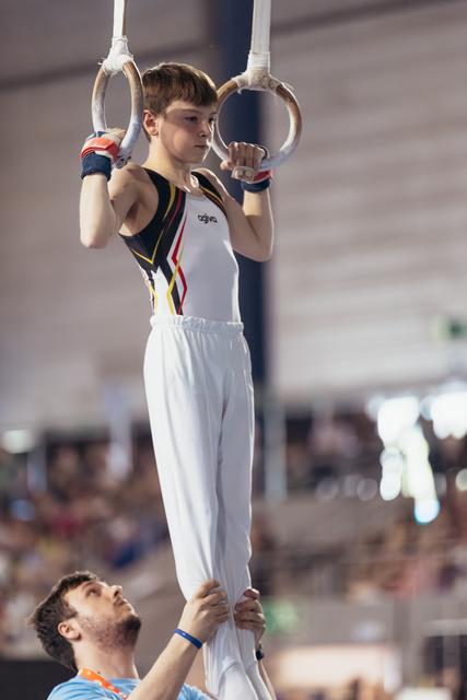 Focused young gymnast grips still rings with determination while coach steadies him from below in a packed arena.