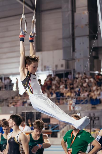 Young male gymnast performs an L-sit on still rings, focused expression, white uniform, crowd watching in background.