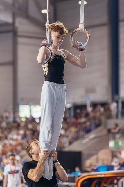 Young male gymnast holds still rings with focused expression while coach steadies his legs in packed gymnastics hall.