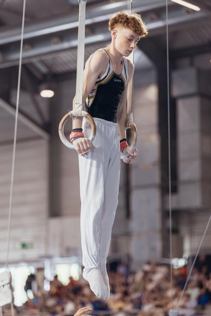 Young male gymnast with red hair grips still rings, looking down with intense focus during a gymnastics competition.