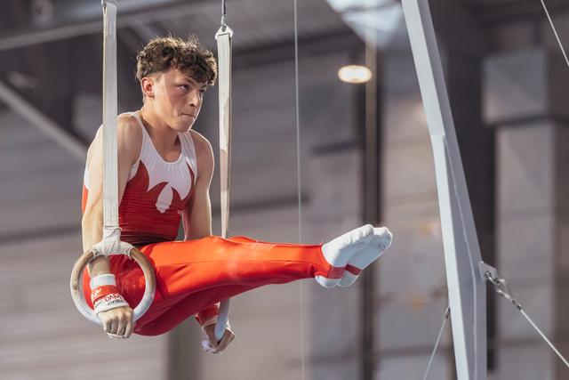 Young male gymnast performs on still rings, legs raised, wearing red uniform, expression of intense concentration.