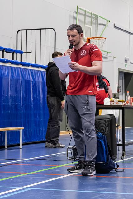 Gespierde man in rood sportshirt leest voor uit papier terwijl hij in een sporthal een microfoon vasthoudt.