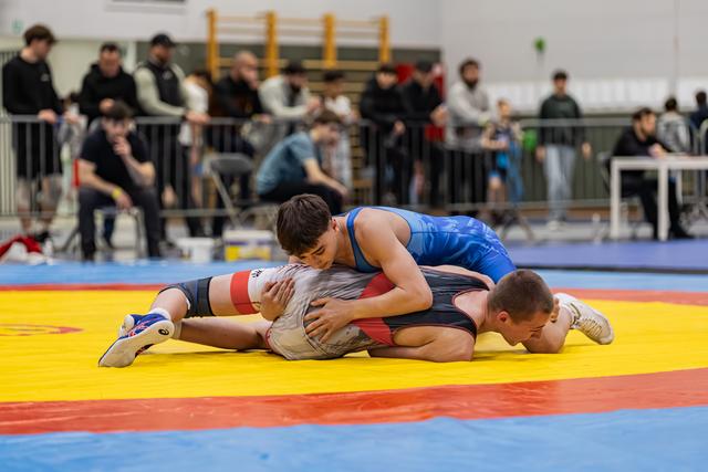 Worstelaar in blauw tenue houdt tegenstander neer op de gele mat tijdens een indoortoernooi.
