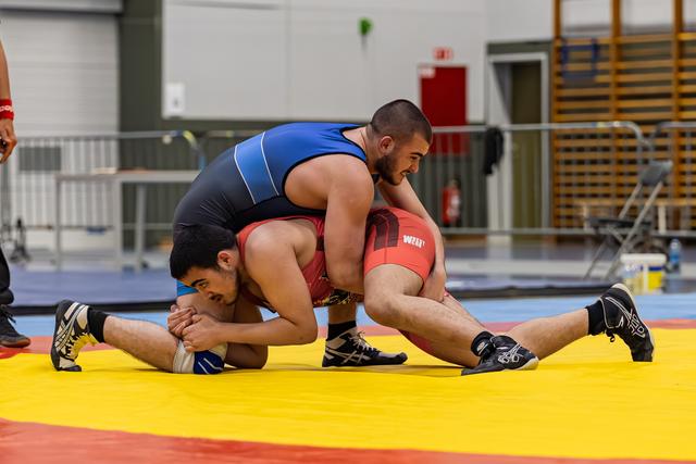 Worstelaar in blauw tenue controleert tegenstander in rood en drukt hem neer op de geel-rode mat.