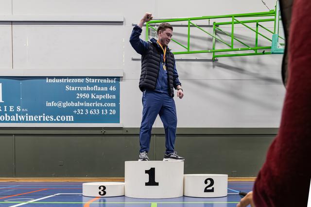 Jonge mannelijke atleet steekt zijn vuist omhoog op het erepodium, met een gouden medaille om zijn nek, breed glimlachend in een sporthal.