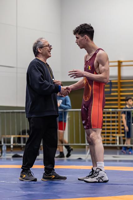 Oudere coach schudt glimlachend de hand van een jonge worstelaar in een bordeauxrood wedstrijdpak op de blauwe mat