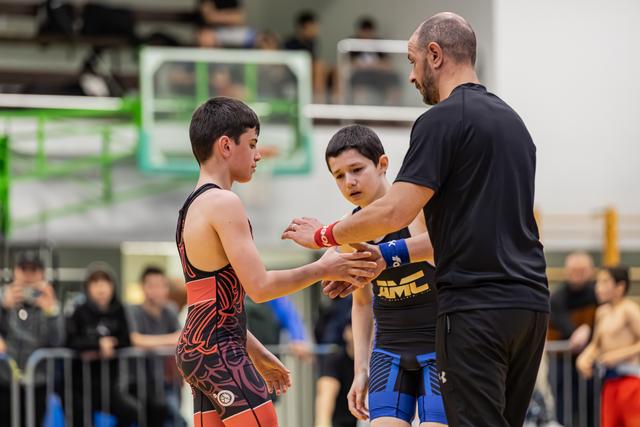 Een coach begeleidt een handdruk tussen twee jonge worstelaars in wedstrijdpak op de mat.