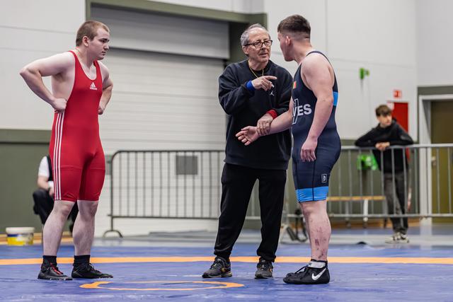 Coach geeft aanwijzingen aan twee worstelaars in rode en blauwe tenue op de mat, beiden luisteren aandachtig.