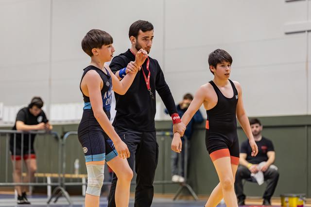 Een coach in het zwart geeft gerichte instructies aan twee jonge worstelaars in een sporthal, één in blauw, één in zwart tenue.